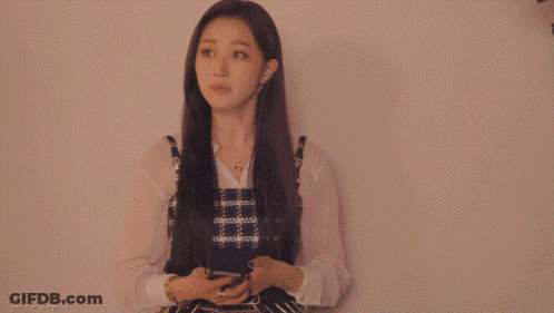 A Woman In A Plaid Dress Is Standing In Front Of A White Wall GIF