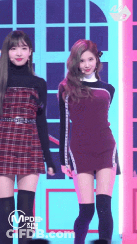A Woman In A Plaid Dress Is Standing Next To Another Woman In A Black Dress GIF
