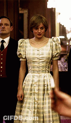A Woman In A Plaid Dress Stands In Front Of A Man In A Suit And Tie GIF