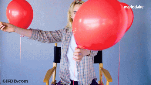 A Woman In A Plaid Shirt Is Holding Two Red Balloons In Front Of Her Face With Marie Claire Written In The Corner GIF
