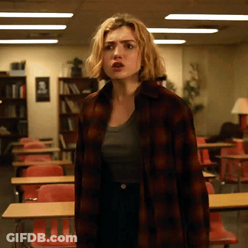 A Woman In A Plaid Shirt Stands In A Classroom With Red Chairs GIF