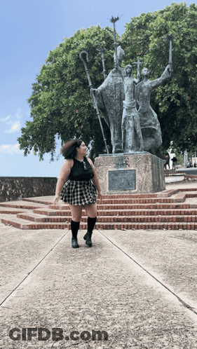 A Woman In A Plaid Skirt Stands In Front Of A Statue Of Three Men GIF