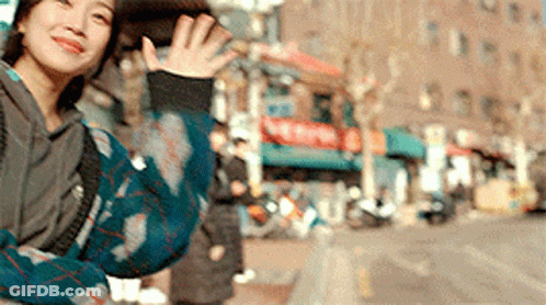 A Woman In A Plaid Sweater Is Waving Her Hand In Front Of A Building . GIF
