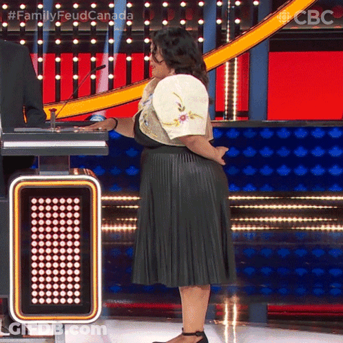 A Woman In A Pleated Skirt Stands On A Stage In Front Of A Sign That Says ' Family Feud Canada ' On It GIF