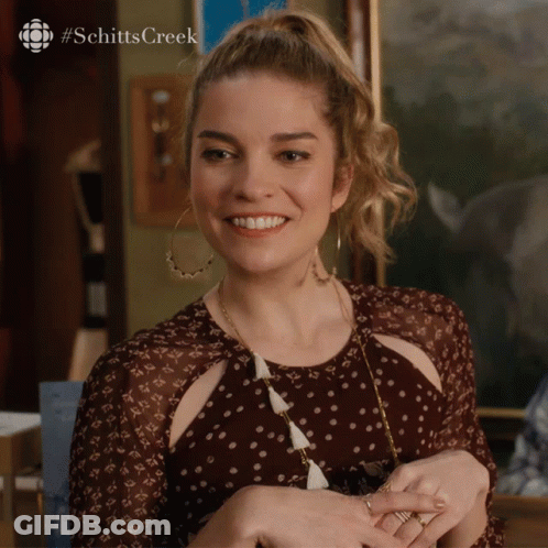 A Woman In A Polka Dot Dress Is Smiling With The #Schittscreek Logo Behind Her GIF