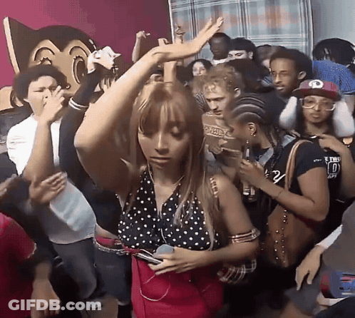 A Woman In A Polka Dot Top Is Dancing In A Crowd With A Man Wearing A Hat With The Letter O On It GIF