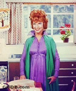 A Woman In A Purple Dress And Green Jacket Stands In Front Of A Window GIF