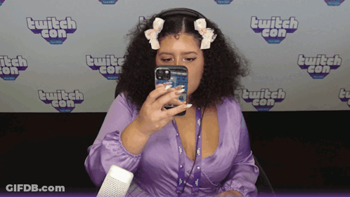 A Woman In A Purple Dress Is Taking A Picture Of Herself In Front Of A Wall That Says Twitch Con GIF