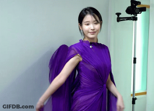 A Woman In A Purple Dress Stands Next To A Camera GIF