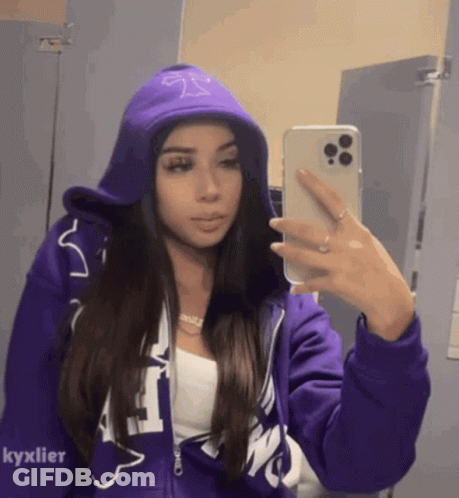 A Woman In A Purple Hoodie Is Taking A Selfie In A Mirror . GIF