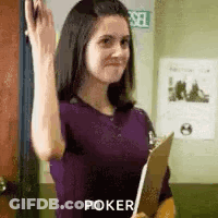 A Woman In A Purple Shirt Is Holding A Clipboard And Waving At The Camera . GIF