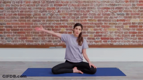 A Woman In A Purple Shirt Sits On A Blue Yoga Mat In Front Of A Brick Wall GIF