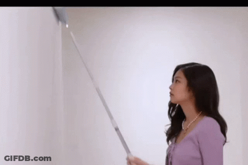 A Woman In A Purple Sweater Is Standing Next To A White Wall And Holding A Vacuum Cleaner . GIF