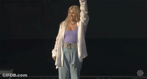 A Woman In A Purple Tank Top And White Shirt Stands On A Stage GIF