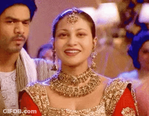 A Woman In A Red And Gold Dress Is Smiling While Standing Next To A Man . GIF