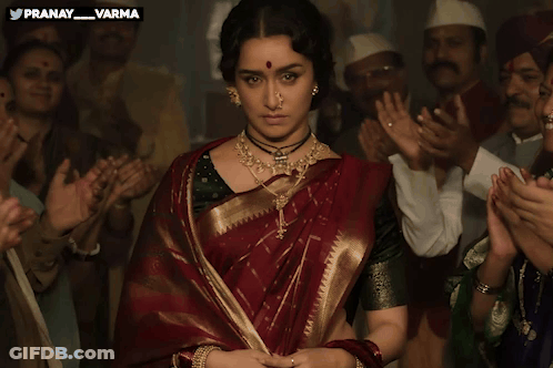 A Woman In A Red And Gold Saree Stands In Front Of A Crowd Of People With The Words Pranay Varma Above Her GIF