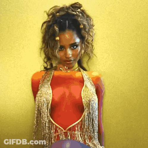 A Woman In A Red And Gold Top With Fringe Is Standing In Front Of A Yellow Wall GIF