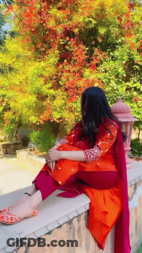A Woman In A Red And Orange Dress Is Sitting On A Wall . GIF