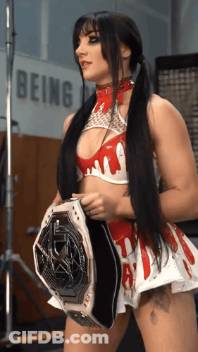 A Woman In A Red And White Outfit Is Holding A Wrestling Belt In Front Of A Sign That Says Being GIF