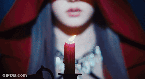 A Woman In A Red Cape Is Holding A Lit Candle In Front Of Her Face GIF