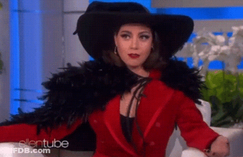 A Woman In A Red Coat And Black Hat Is Sitting On A Chair . GIF