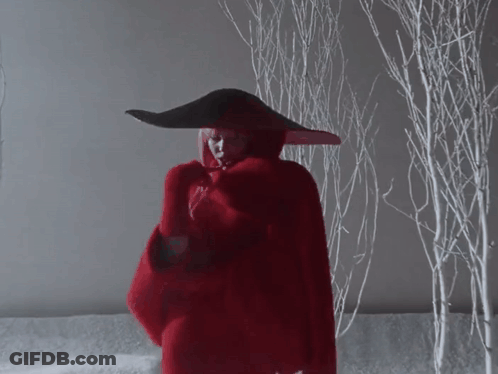 A Woman In A Red Coat And Black Hat Stands In Front Of Snowy Trees GIF