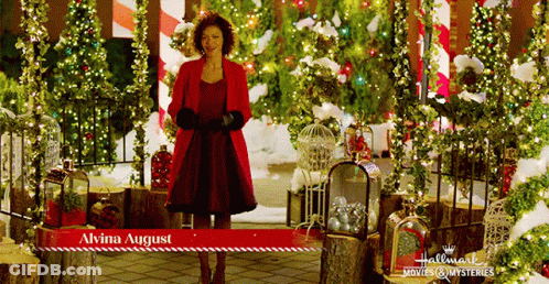 A Woman In A Red Coat Is Standing In Front Of Christmas Decorations And A Sign That Says " Alvina August " GIF