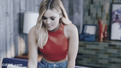 A Woman In A Red Crop Top And Blue Jeans Is Sitting On A Couch GIF