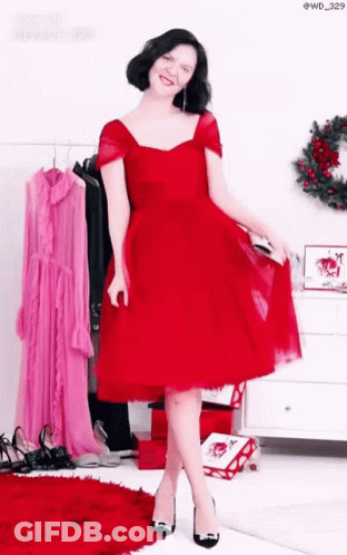 A Woman In A Red Dress And Black Heels Is Standing In Front Of A Wreath . GIF