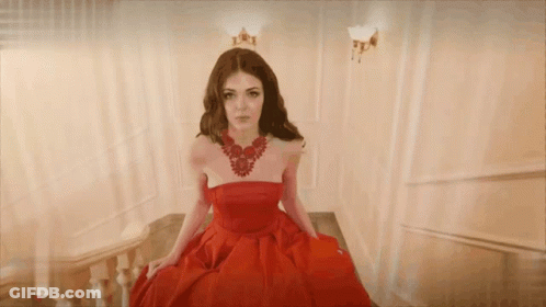 A Woman In A Red Dress And Necklace Is Sitting On A Set Of Stairs GIF