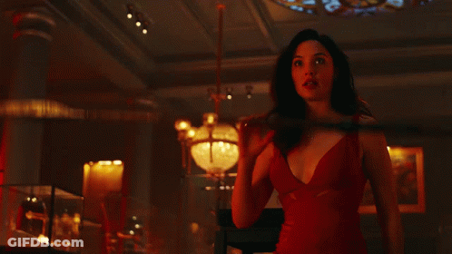 A Woman In A Red Dress Is Holding A Spear In A Dark Room GIF