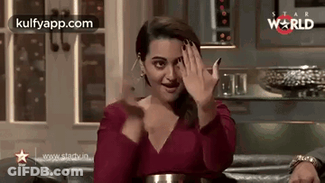 A Woman In A Red Dress Is Sitting On A Couch And Waving Her Hands . GIF
