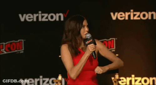 A Woman In A Red Dress Is Speaking Into A Microphone In Front Of A Verizon Sign GIF