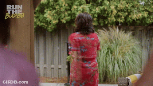 A Woman In A Red Dress Is Standing In Front Of A Fence With The Words Run The Burbs On The Bottom GIF
