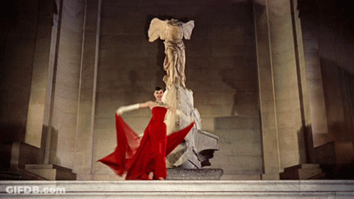 A Woman In A Red Dress Is Standing In Front Of A Large Statue GIF