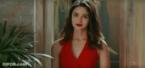 A Woman In A Red Dress Is Standing In Front Of A Plant And Looking At The Camera . GIF