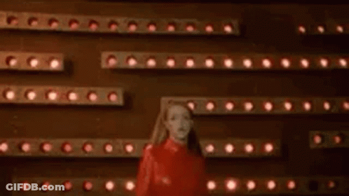 A Woman In A Red Dress Is Standing In Front Of A Wall Of Lights And A Sign That Says Oops . GIF