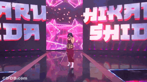 A Woman In A Red Dress Is Standing On A Stage In Front Of A Large Screen That Says ' Nikki Shida ' GIF