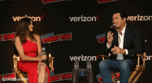 A Woman In A Red Dress Sits Next To A Man Holding A Microphone At A Comic Con GIF