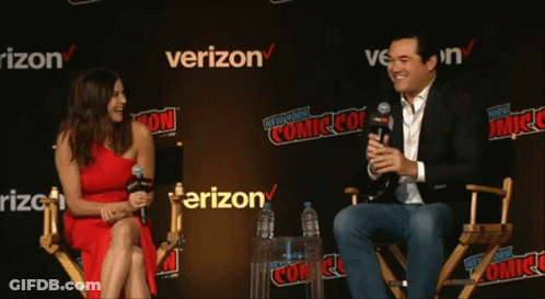 A Woman In A Red Dress Sits Next To A Man In A Suit In Front Of A Verizon Sign GIF