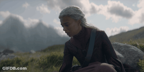 A Woman In A Red Dress Sits On A Rock In A Field With Mountains In The Background GIF