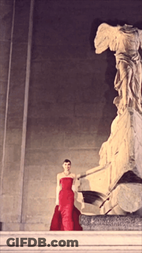 A Woman In A Red Dress Stands Next To A Statue GIF