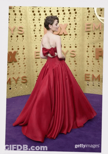 A Woman In A Red Dress Stands On A Purple Carpet In Front Of A Wall That Says Emmys GIF