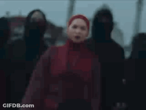 A Woman In A Red Hijab Is Standing In Front Of A Group Of People In Black Hoods . GIF