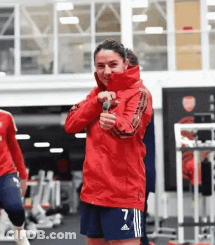 A Woman In A Red Jacket And Blue Shorts Is Standing In A Gym Holding A Microphone . GIF