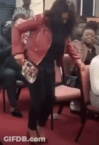 A Woman In A Red Jacket Is Dancing In Front Of A Crowd Of People Sitting In Chairs . GIF