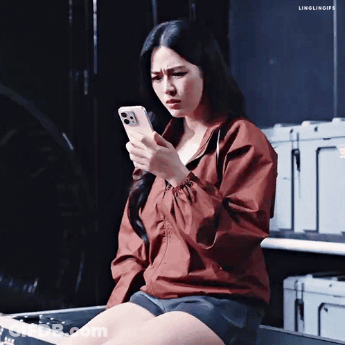 A Woman In A Red Jacket Is Sitting On A Box Looking At Her Phone GIF