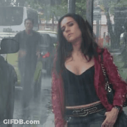 A Woman In A Red Jacket Is Standing In The Rain In Front Of A Bus Stop . GIF