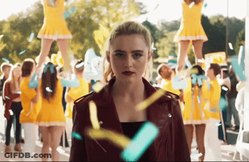 A Woman In A Red Jacket Stands In Front Of Cheerleaders GIF