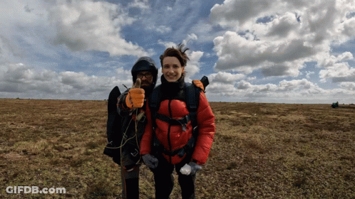 A Woman In A Red Jacket Stands Next To A Man In A Black Jacket Giving A Thumbs Up GIF